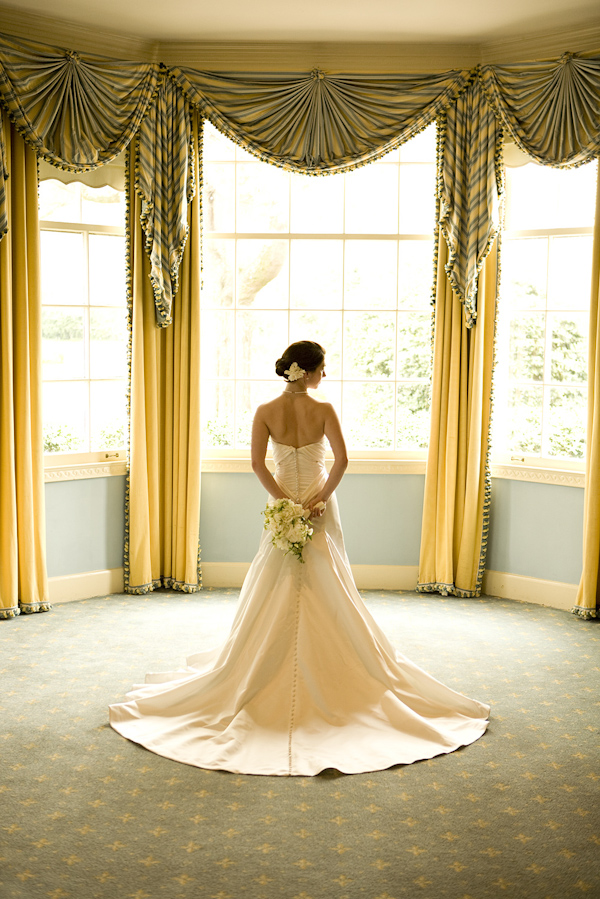 bride standing in front of beautiful bay windows in ivory a-line dress wearing a ivory floral hairpiece and holding white and green bouquet behind her back - photo by North Carolina based wedding photographers Cunningham Photo Artists
